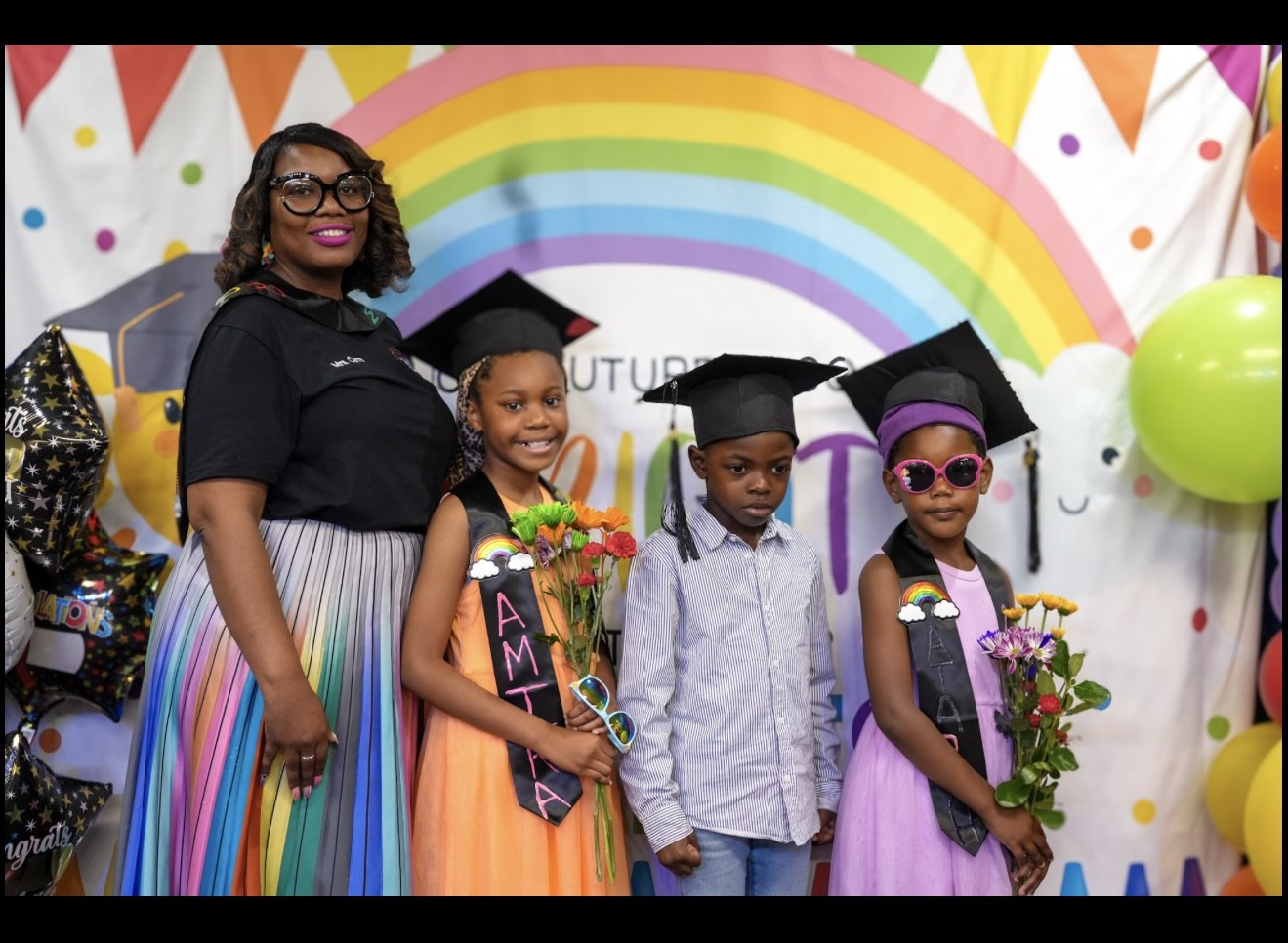An Academy staff member with graduates beside the rainbow graduation backdrop